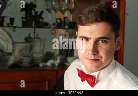 Ritratto di un giovane con una camicia bianca e un papillon rosso. Bell'uomo seduto in un ambiente interno con sfondo vintage sfocato. Uomo Foto Stock