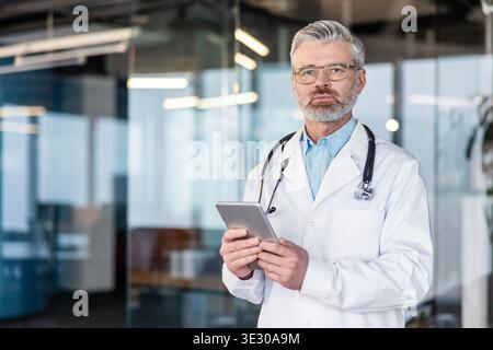 Adulto professionista sanitario maschile che indossa un camice da laboratorio e uno stetoscopio, tenendo una compressa digitale mentre si trova in una moderna clinica medica o corridoio ospedaliero Foto Stock