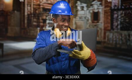 Operaio edile nero con elmetto e tute blu che tengono il computer portatile e scrivono con i guanti; concentratevi sulla produttività. Foto Stock