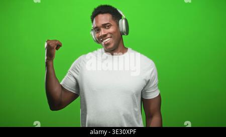 L'uomo nero che indossa le cuffie si flette bicipite destro e sorride verso la fotocamera in uno studio green screen indossando una t shirt bianca; un ritmo sicuro. Foto Stock