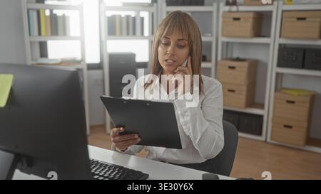 La donna tiene gli appunti, il telefono all'orecchio mentre legge i documenti alla scrivania dell'edificio vicino al monitor e ai file impilati; documenti sulla concentrazione. Foto Stock