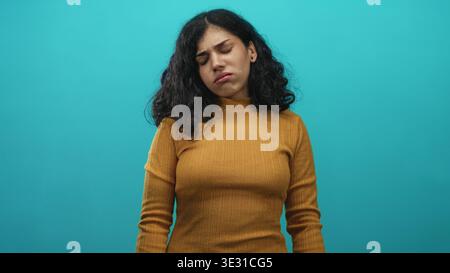 Giovane donna araba faccia con occhi chiusi ed espressione dolorosa con maglione senape in studio turchese, leggera inclinazione della testa e sguardo basso; tristezza Foto Stock