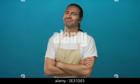 Uomo biondo con i capelli lunghi che indossa il grembiule attraversa le braccia e sorride a occhi chiusi in studio blu; serenità. Foto Stock