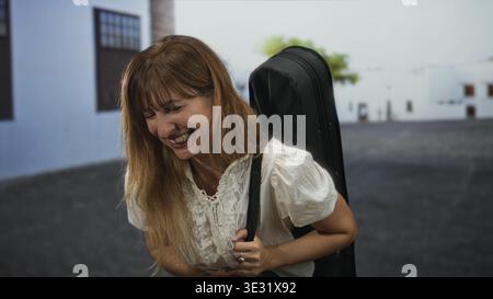 Donna che ride e tiene in mano una tracolla per chitarra su una strada illuminata dal sole con edifici bianchi e una palma; viaggio divertente. Foto Stock