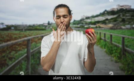 Uomo che tiene la mela in mano destra, dita a labbra su un sentiero sul lungomare con camicia bianca e barba, collina del villaggio in lontananza; calma consapevole. Foto Stock