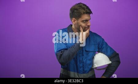 Uomo operaio edile in tute da lavoro blu con elmetto bianco con mano sul mento e sguardo concentrato sullo sfondo viola dello studio; focu premuroso Foto Stock