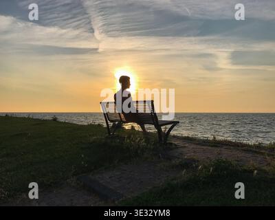 Silhouette di un uomo seduto su una panchina, che guarda tranquillamente un tramonto dorato sul mare. La luce calda si riflette sull'acqua, creando un'atmosfera tranquilla. Foto Stock