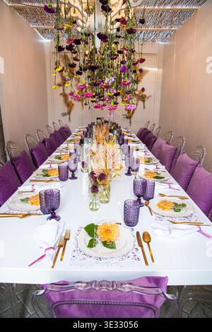 Tavolo per ricevimenti nuziali splendidamente arredato con bicchieri in tessuto viola, posate in oro e delicati centrotavola floreali. Foto Stock