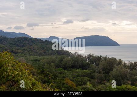 Isola di Koh Tarutao con vegetazione lussureggiante e montagne lontane. Il mare calmo incontra la costa sotto un cielo nuvoloso. Foto Stock