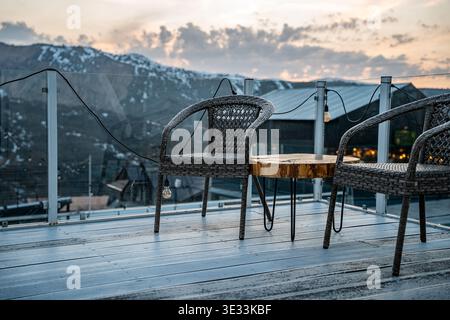 Un'area salotto all'aperto tranquilla con due sedie in vimini e un tavolo in legno, adagiata sullo sfondo di montagne e di uno splendido cielo al tramonto. Foto Stock