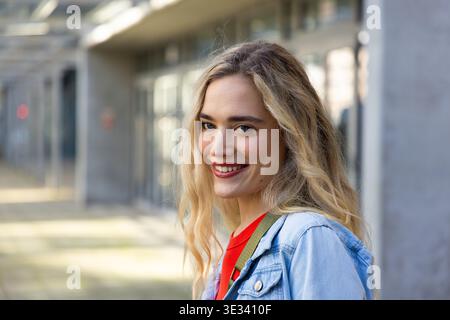 Donna sorridente, in piedi sul vialetto lastricato dell'edificio, con giacca in denim e top rosso, spazio copia Foto Stock