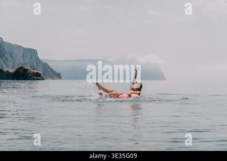 Donna che nuota spruzza giovane donna che galleggia su un anello gonfiabile in mare facendo spruzzi d'acqua con montagne panoramiche alle spalle Foto Stock