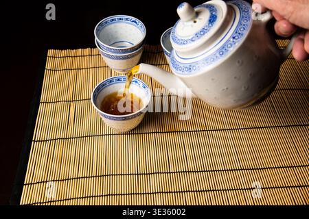 Il tradizionale tè cinese viene versato da una teiera in una tazza, esposto su un tappetino di bambù, mettendo in risalto la cultura del tè asiatico e la presentazione serena. Foto Stock