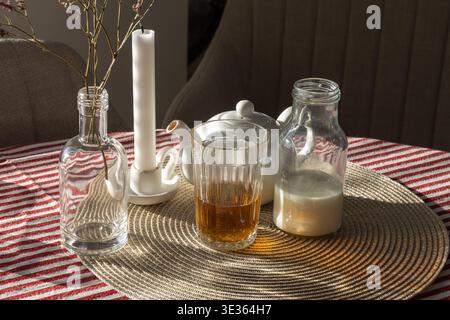 Bicchiere di tè e bottiglia di latte disposti su un tappeto intrecciato con teiera bianca e candela. La luce calda del sole crea ombre morbide e un'accogliente scena di natura morta in una Foto Stock