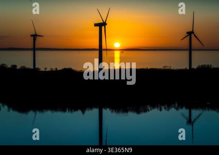 Le turbine eoliche si stagliano contro il tramonto sull'acqua con riflessi su una superficie calma. Foto Stock