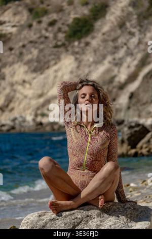 Donna, spiaggia, oceano, siede sulla roccia vicino al mare, rilassandosi pacificamente sotto il sole Foto Stock
