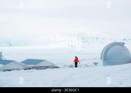 Osservatorio Mount John, nuova Zelanda: Una donna solitaria in giacca arancione cammina nella neve profonda vicino a una cupola del telescopio astronomico. Foto Stock