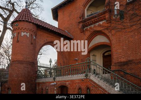 Uno storico edificio in mattoni rossi caratterizzato da un passaggio ad arco, scalinata in pietra, ringhiera decorativa in ferro e una torre rotonda con tetto piastrellato. Dettagliato Foto Stock
