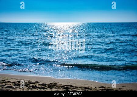 Il sole splende brillantemente su un mare calmo, creando riflessi dorati sulle acque tranquille. Foto Stock