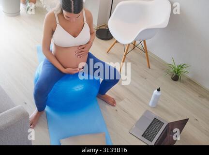 Aspettandosi che la madre faccia esercizi di gravidanza su una palla blu nel suo salotto per uno stile di vita sano Foto Stock