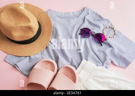 Outfit estivo casual con cappello, occhiali da sole, T-shirt blu, pantaloncini bianchi e sandali su sfondo rosa. Composizione minimale in stile piatto. Vista dall'alto. Foto Stock