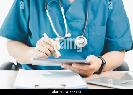 Medico in scrub blu utilizzando tablet e stilo con stetoscopio, documentando le cartelle dei pazienti al banco della clinica, concetto di salute digitale, t Foto Stock