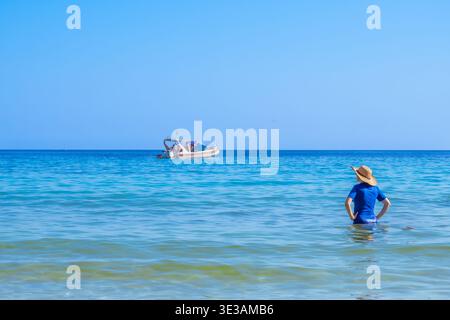 Una giovane donna con costume da bagno blu e cappello di paglia si staglia con la parte superiore del corpo nelle acque turchesi del mare, guardando un motoscafo con turisti Foto Stock