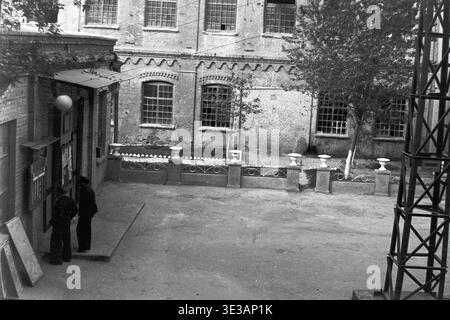 Fotografia vintage degli anni '1950-'1960 del cortile dello stabilimento AIZ a Slavyansk. Due operai di sesso maschile in uniforme scura si trovano vicino all'ingresso di un edificio di fabbrica in mattoni con grandi finestre a più pannelli. Sulla destra è visibile una torre reticolare metallica con un cartello che riporta la linea n. 1. Piccoli vasi decorativi fiancheggiano il passaggio pedonale. Un assaggio della storia industriale e della vita lavorativa quotidiana prima della guerra, mostrando i dettagli architettonici e l'atmosfera di lavoro di un impianto di produzione sovietico nell'era di un tranquillo Donbas. Foto Stock