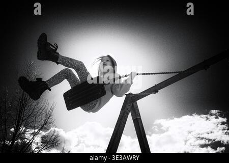 Foto in bianco e nero del bambino che oscilla in alto contro il cielo luminoso con una forte luce solare. Scena dinamica ed emotiva che simboleggia libertà, gioia e carefre Foto Stock