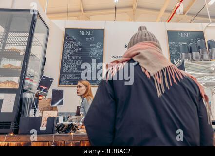 Una giovane donna caucasica con capelli rossi serve caffè in un caffè. Un uomo di mezza età con un cappello e una sciarpa si trova al bancone, guardando il menu. Foto Stock