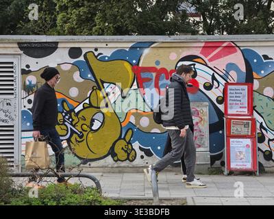 Persone che camminano lungo un muro di graffiti a Monaco, vibrante scena urbana con pedoni che passano attraverso pittoreschi murali di Street art, scritte audaci ed expres Foto Stock