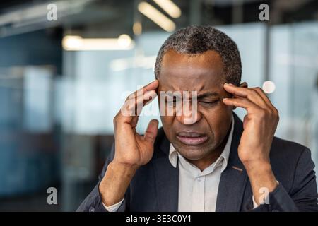 Uomo d'affari maturo che soffre di mal di testa e disagio intenso mentre lavora in remoto nel suo ufficio moderno, mostrando segni di dolore e stress travolgente da lavoro impegnativo Foto Stock