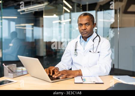 Medico maschile che indossa un camice da laboratorio bianco e uno stetoscopio, guardando direttamente la fotocamera mentre digita su un computer portatile, fornendo servizi sanitari in un ambiente professionale di ufficio Foto Stock