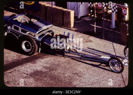 Vista rialzata di un dragster anteriore e di un veicolo trainante nell'area del paddock durante la NHRA Winternationals 1967, Pomona, California, febbraio 1967. Il telaio lungo e il motore esposto riflettono il design dei migliori dragster a gas e a combustibile della fine degli anni '1960. Foto Stock