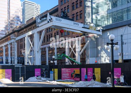 La grande gru a cavalletto bianca si estende su un cantiere del centro tra gli edifici, con la neve a terra e i pedoni che camminano nelle vicinanze. Foto Stock