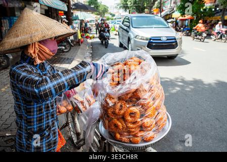 Il venditore ambulante in cappello conico tiene un vassoio di impasto fritto dorato. Indossa maschera facciale e camicia a quadri mentre è in piedi accanto alla bicicletta carica di foo Foto Stock