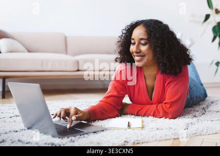 Donna che lavora su un notebook mentre è sdraiata sul pavimento in un accogliente salotto Foto Stock