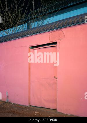 Una parete dipinta di rosa brillante con una porta in metallo chiusa e un tradizionale tetto piastrellato illuminato in una strada buia di notte. La scena rappresenta il contr. Urbano Foto Stock