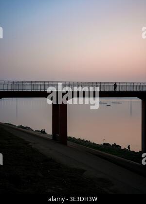 Una figura solitaria sagomata che cammina su un molo pedonale sopraelevato sopra un fiume calmo che riflette il morbido cielo crepuscolo con edifici distanti. La scena Foto Stock