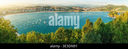 Vista panoramica della baia di la Concha a San Sebastián con spiaggia sabbiosa, yacht e barche sul mare calmo e sulla costa della città circondata da verdi colline a. Foto Stock
