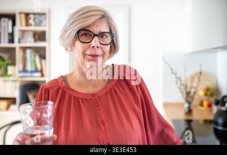 Donna anziana che tiene un bicchiere d'acqua a casa in cucina. Foto Stock