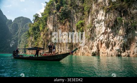 Lago Cheow LAN, Thailandia – 19 febbraio 2020: I turisti si trovano sulla prua di una barca a coda lunga sulle acque turchesi accanto alle scogliere calcaree. Foto Stock