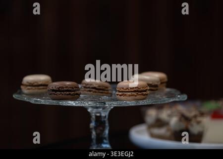 Macaron francesi serviti su un banco di vetro in un ambiente da caffe' con luce soffusa e profondità di campo bassa. Elegante presentazione di dessert con pasticceria di alta qualità Foto Stock