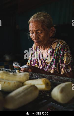 Balikpapan, Indonesia - 15 febbraio 2026. Un ritratto di una donna anziana che si concentra sulla preparazione del Pisang Gapit, un tradizionale spuntino grigliato alla banana. Foto Stock