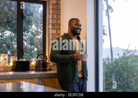 Uomo afroamericano che guarda fuori dalla finestra di vetro in cucina indossando una camicia verde e tenendo in mano una tazza di ceramica Foto Stock