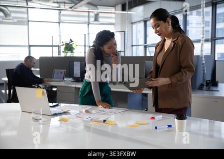 Diverse colleghe che si appoggiano al tavolo indossando abiti professionali, esaminando grafici e tablet Foto Stock