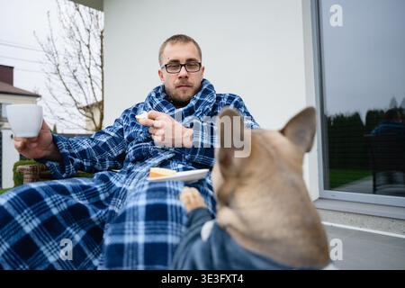 Un uomo in abito a quadri siede sul patio con un caffè e nutre il suo bulldog francese. Routine mattutina tranquilla a casa, momenti di vita intimi con gli animali domestici. Foto Stock