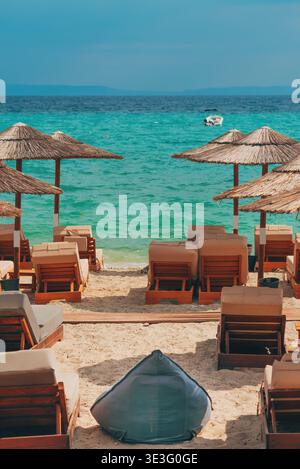 Spiaggia sabbiosa con sedie a sdraio e ombrelloni di paglia di fronte al mare turchese. Rilassante paesaggio costiero con acque calme e barca lontana sotto un cielo limpido. Io Foto Stock
