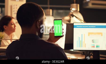 Il dipendente che si occupa dell'avvio tiene in mano uno smartphone con schermo verde e lavora sui dati aziendali, guardando il display mockup isolato. Utilizzo di spazio di copia della chiave cromatica vuota sul modello del telefono cellulare. Foto Stock