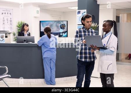 Giovane uomo asiatico discutere diagnosi e trattamento con medico donna in sala d'attesa ospedale. Medico nero con laptop esamina le cartelle cliniche mentre parla con un paziente di sesso maschile nella hall della clinica. Foto Stock